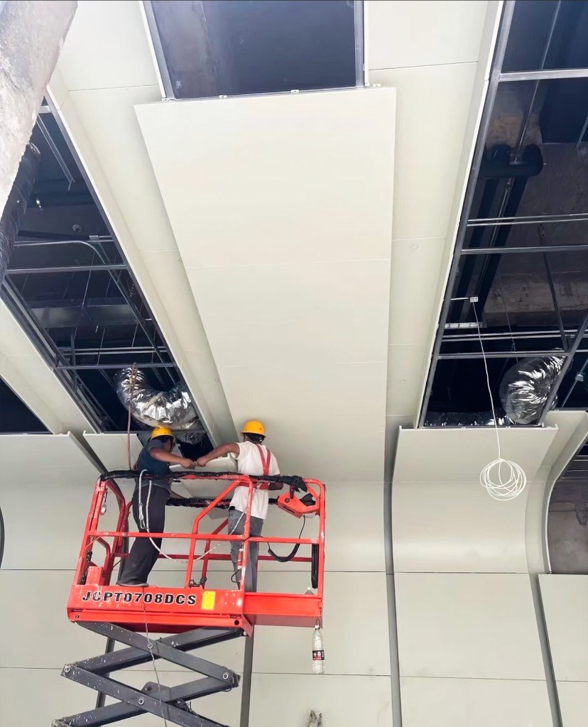 Installers on a scissor lift setting large-format LINDELACR metal ceiling trays to a coordinated grid, ducts still open in the plenum