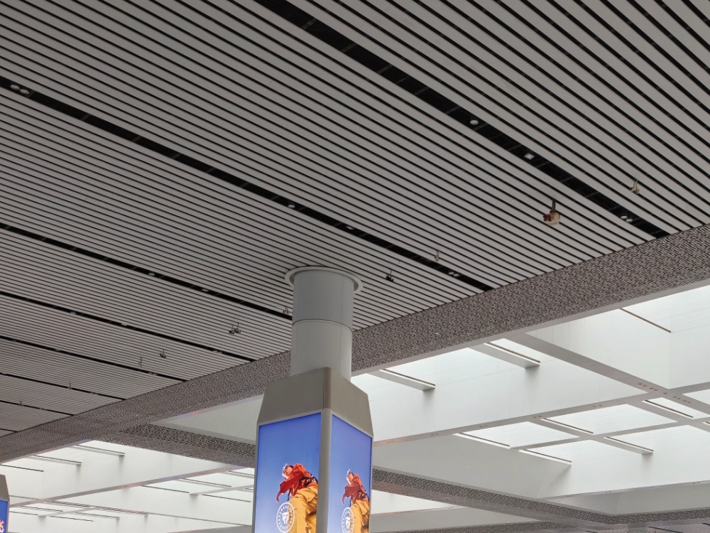 LINDELACR white square-tube baffle ceiling in a public hall: open gaps between slats, column wrap, and a lay-in tray ceiling beyond