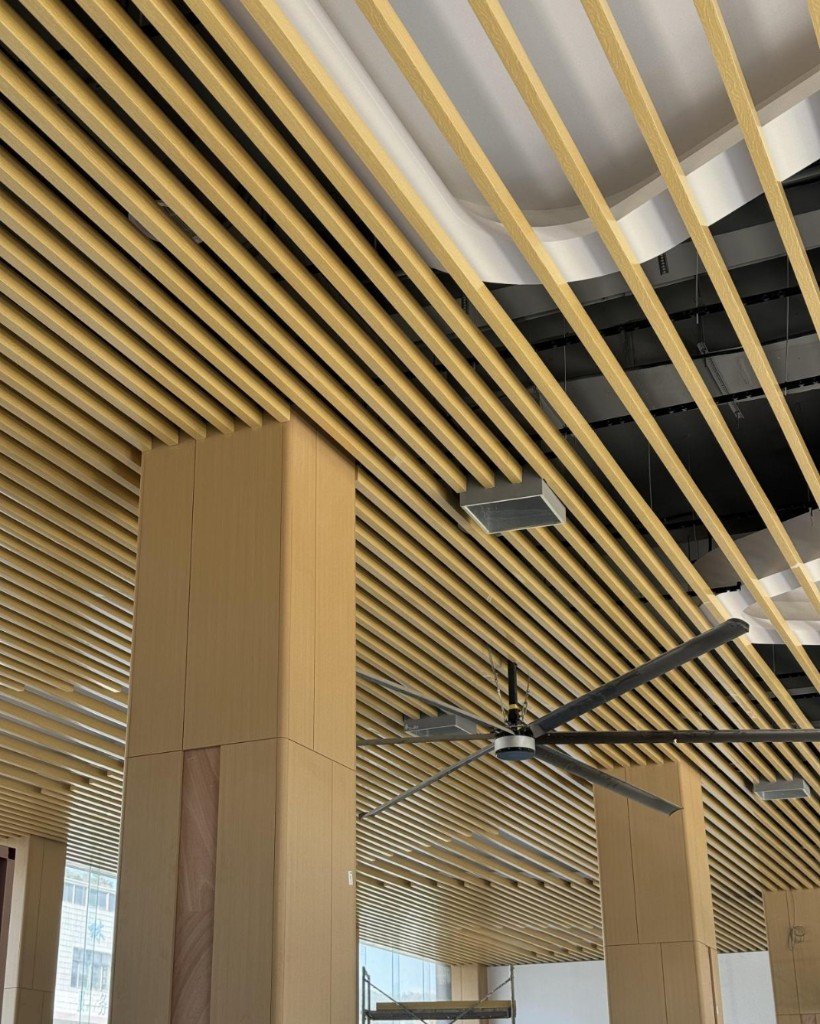Wood-grain linear aluminum baffles over a hall, with a ceiling fan and ducts still visible in the open plenum
